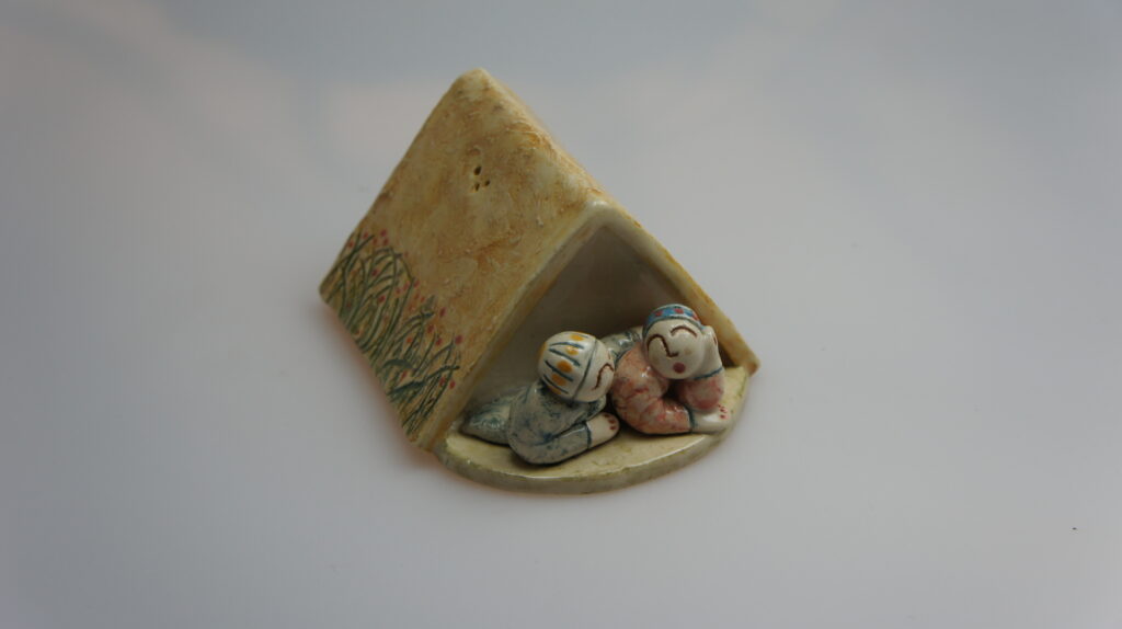 Een keramisch peper- en zoutstel met het thema 'Samen Kamperen', ontworpen door de getalenteerde kunstenares Ank Uitenbroek. Het stel toont een schilderachtige scène van een tent met twee mensen ervoor, omringd door gras, en de ene persoon slaat teder een arm om de ander.